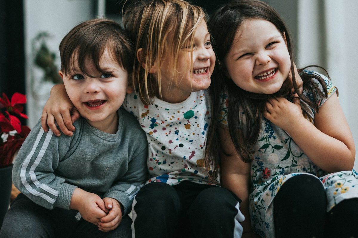 Three young children sitting close together on front steps, laughing and hugging